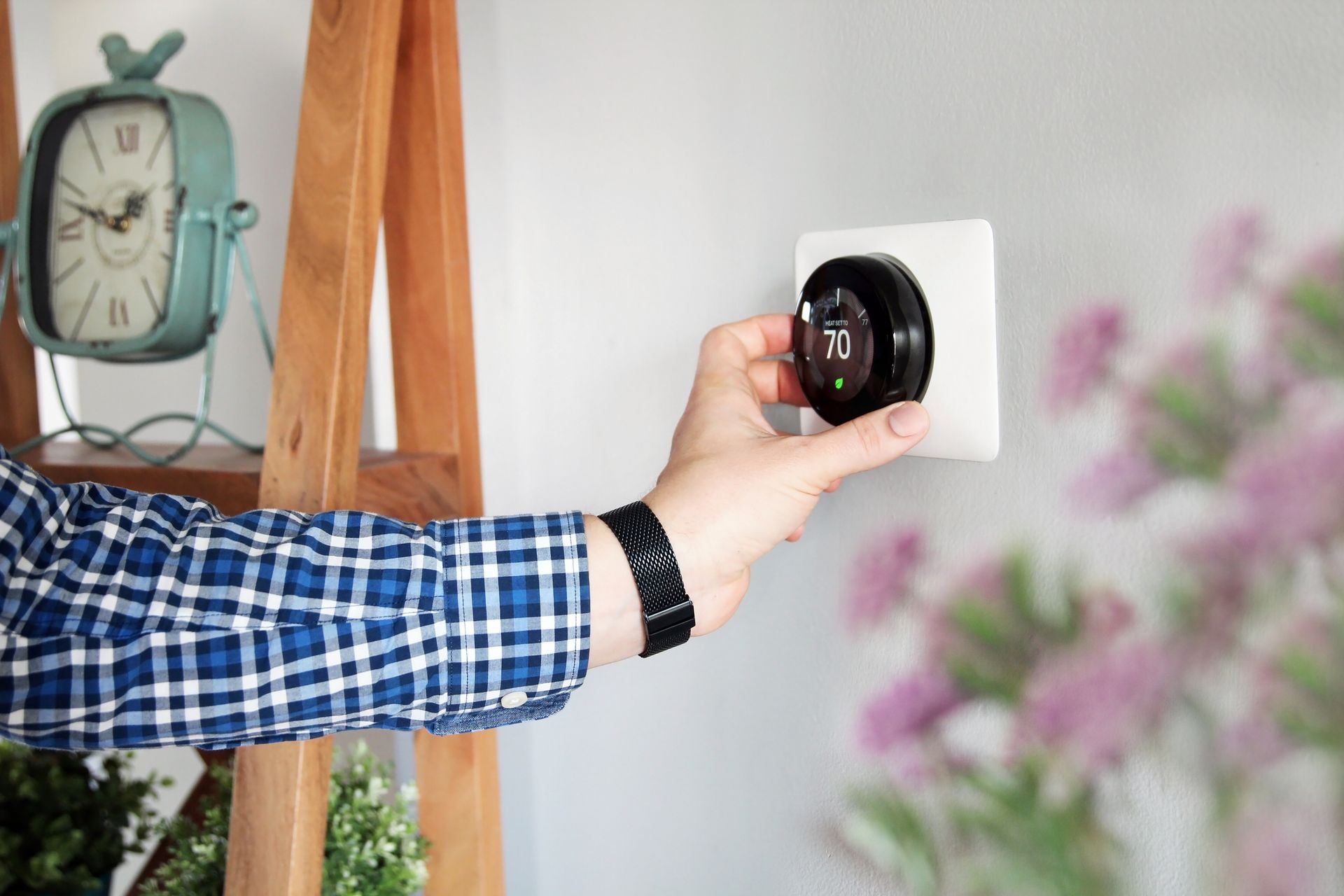 A hand in a blue plaid shirt adjusts a circular smart thermostat on a white wall near a wooden shelf and a clock.