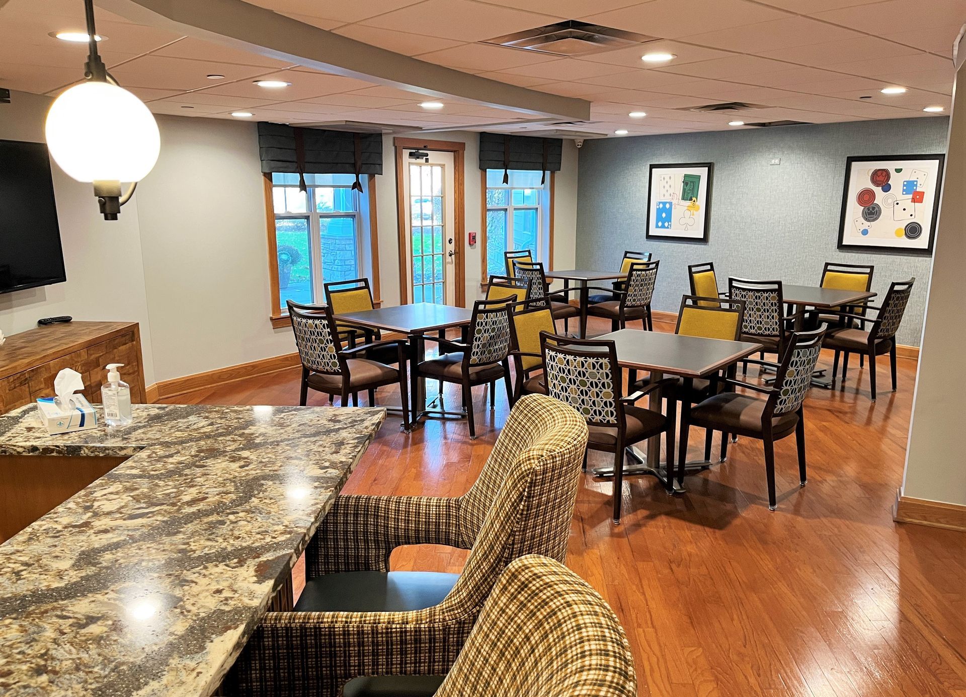 A dining area with tables, chairs, and a bar with stools.  Wooden floors, framed artwork, and large windows.