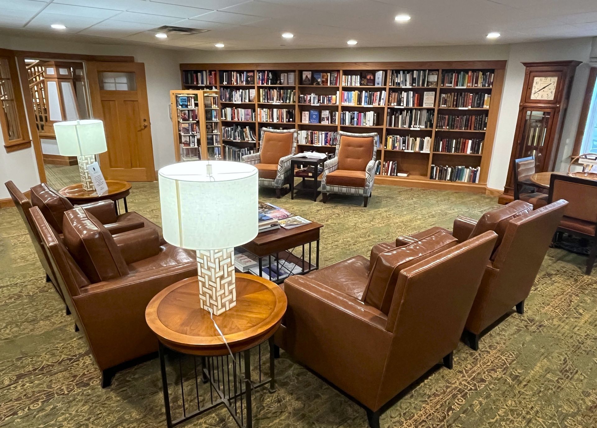 A cozy library seating area with brown leather armchairs arranged in a circle around lamps and a small table. Bookshelves and a grandfather clock are in the background.