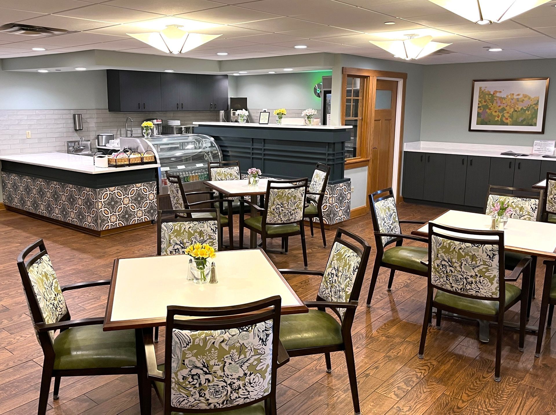 A dining room with square tables and chairs, and a buffet area. The chairs are green with floral patterns, and the walls are gray.