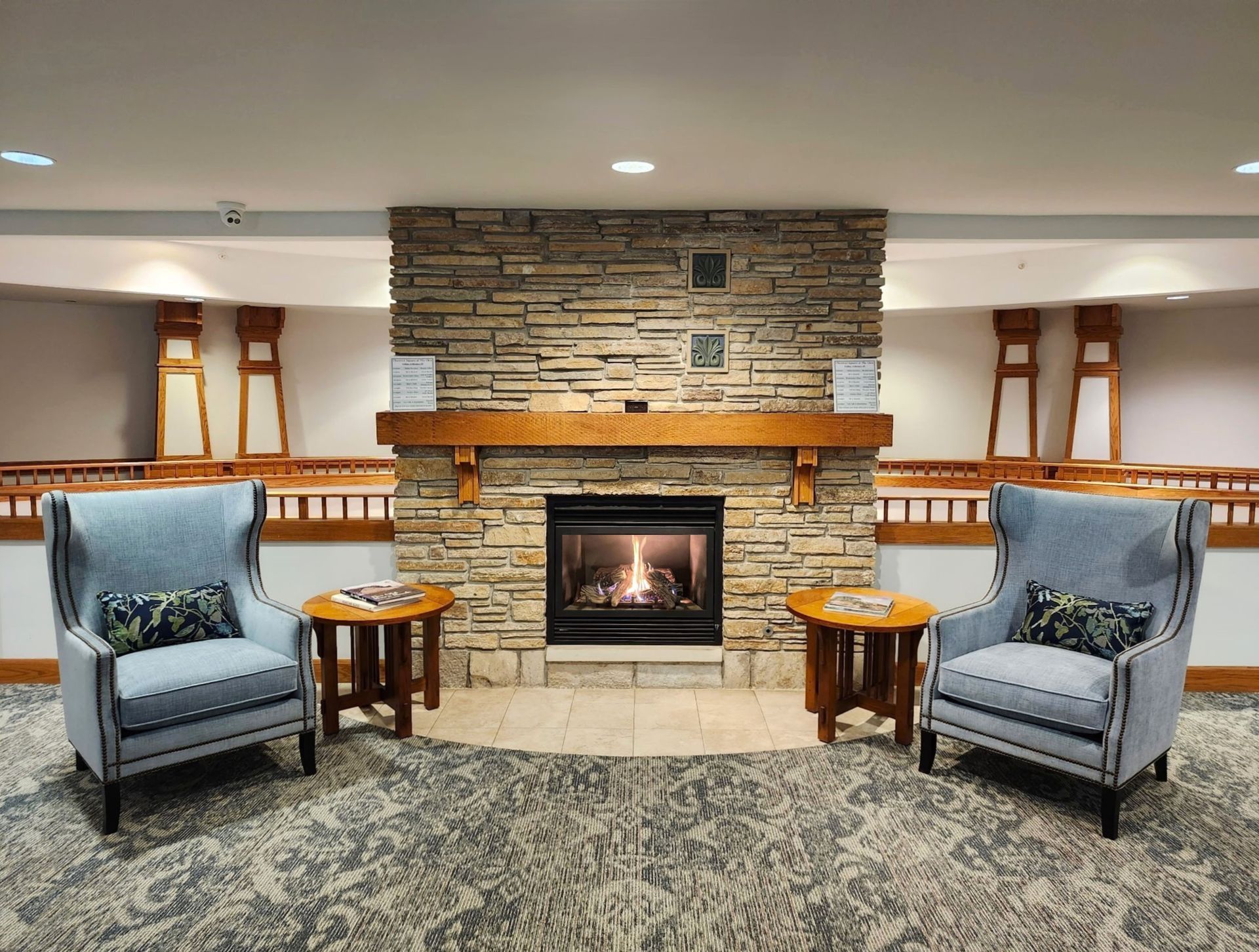 Two blue armchairs flank a stone fireplace with a lit fire, small wooden tables beside them, and a patterned carpet in a waiting area.