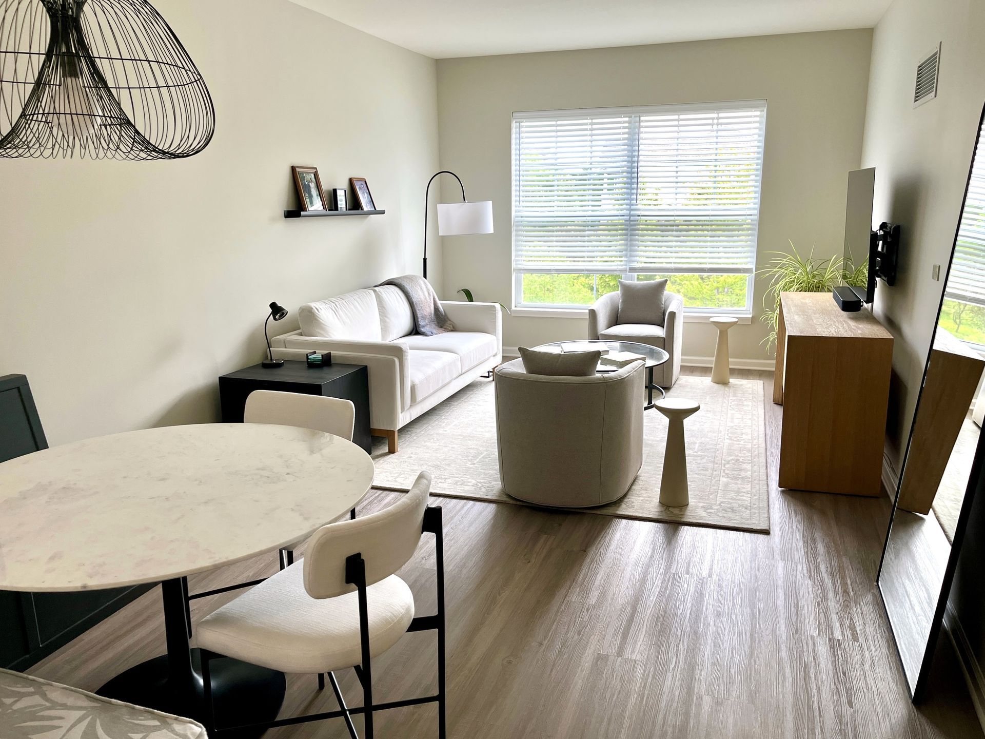 Small, modern living room with a white sofa, round dining table, and a large window. Neutral tones and natural light.