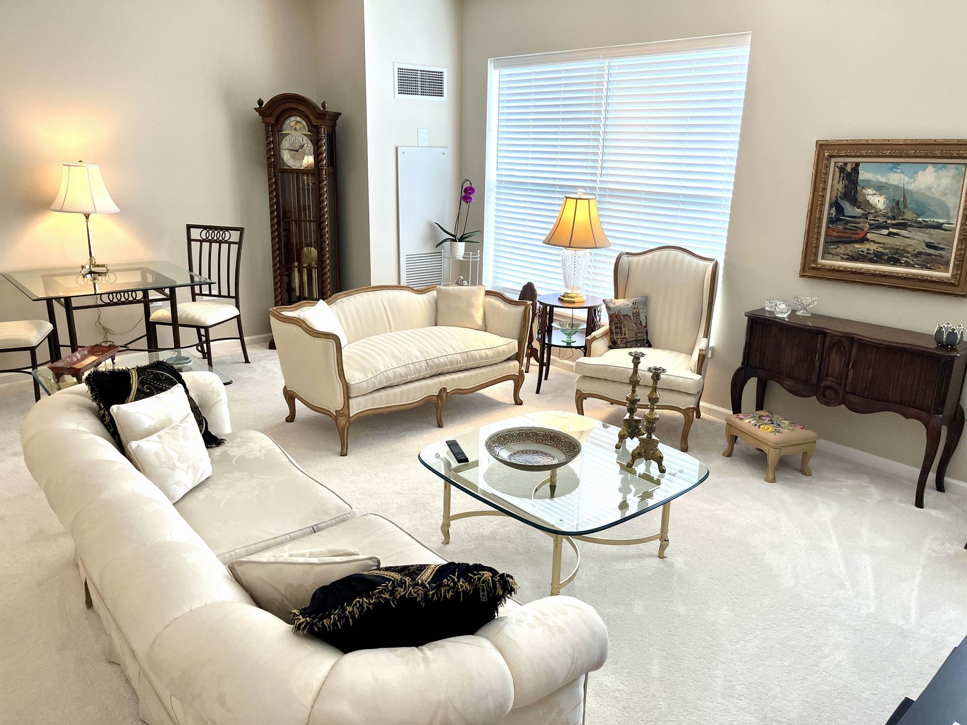 Living room with light-colored furniture, including a sofa, chairs, and a grandfather clock. A coffee table and lamps are also present.