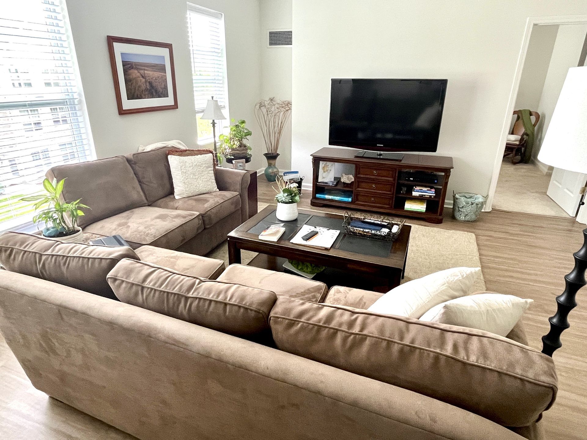 A living room with a brown sectional sofa, TV, coffee table, and artwork. Natural light floods the space.