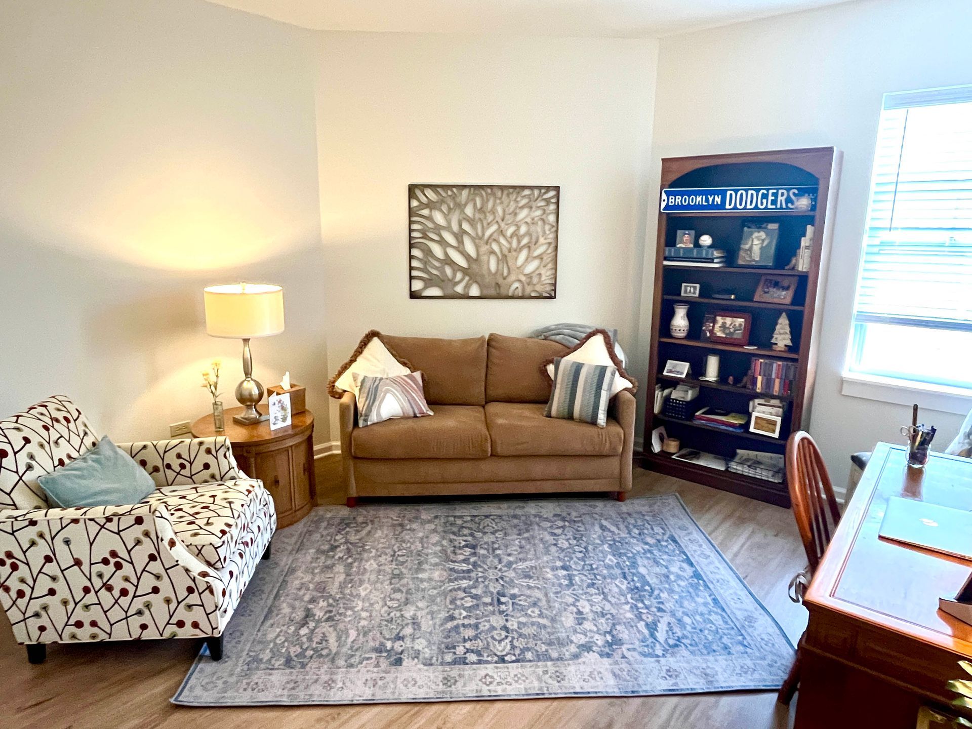 Cozy home office with a brown sofa, patterned armchair, bookshelf, desk, and rug.