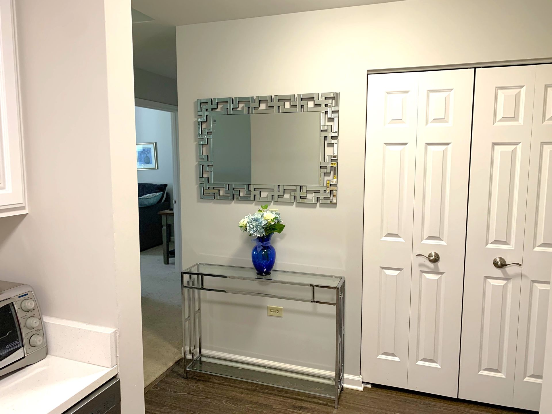 A small foyer with a decorative mirror, glass table, blue vase with flowers, and a white closet.