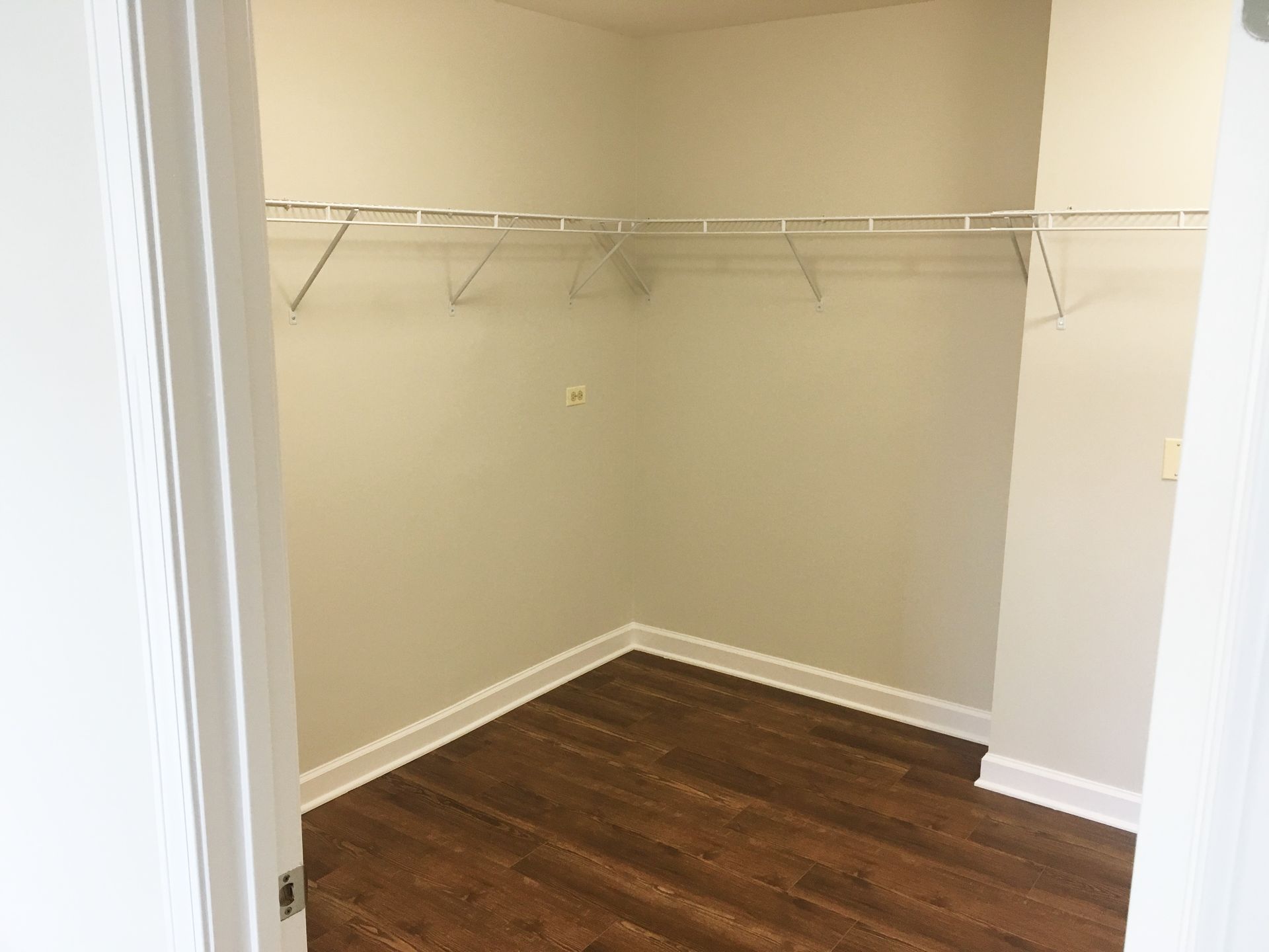 An empty walk-in closet with a wire shelf. It has brown flooring and light gray walls, framed by a white doorway.
