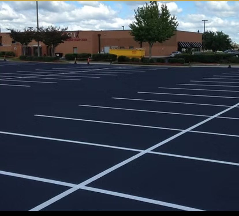 A worker in an orange jumpsuit uses a machine to paint a white line on a black asphalt road.