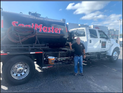 A worker uses a squeegee to apply black asphalt sealer to a driveway, with several buckets of sealant nearby.
