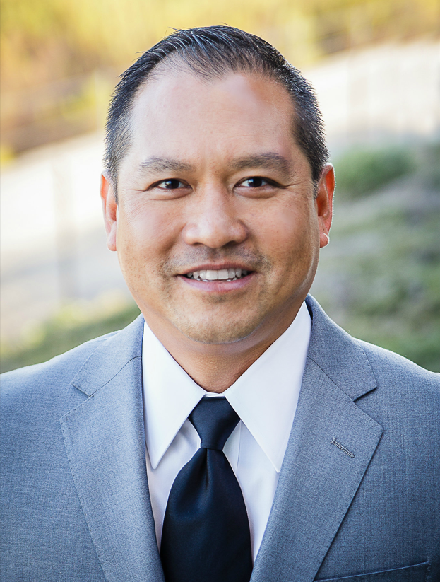 Man in light blue checkered shirt smiling, leaning against a building.