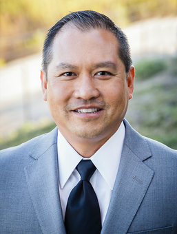 Man in gray suit and black tie smiling at the camera. Outdoor setting.