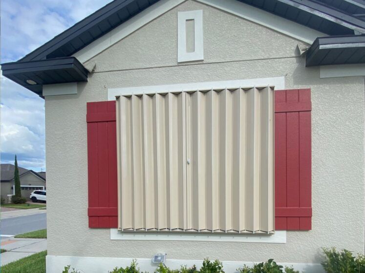 House with tan accordion shutters and red shutters; cloudy sky.