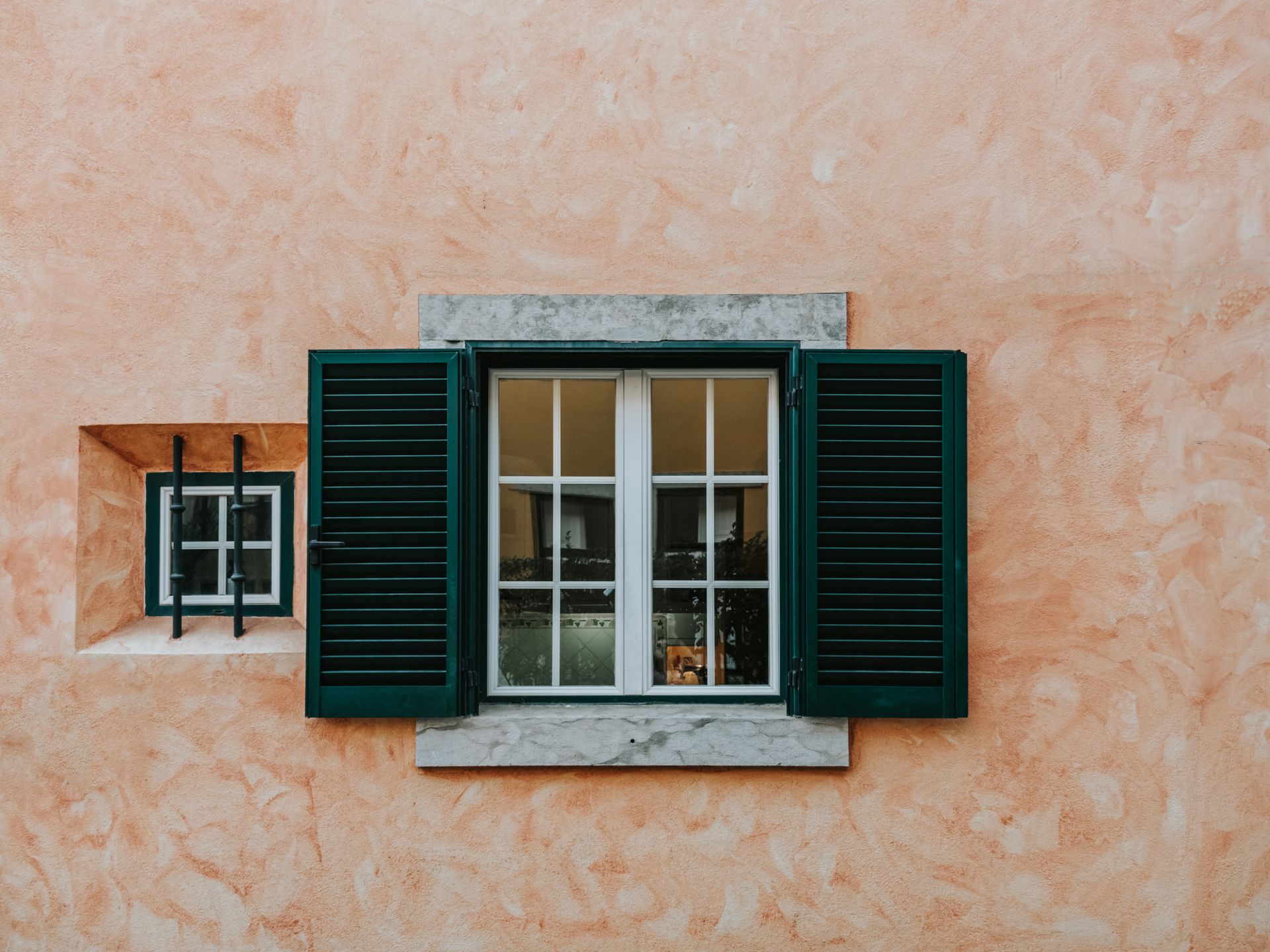 Peach-colored wall with two windows; one with green shutters, the other with a small window and black bars.
