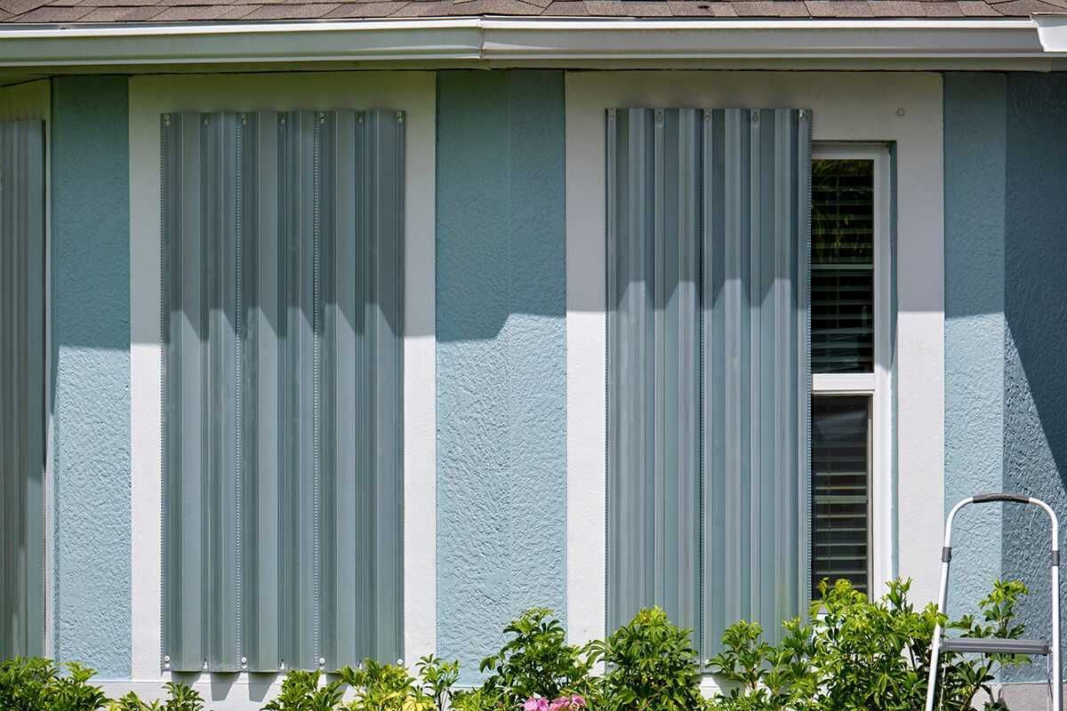 Two windows with corrugated metal storm shutters, next to light blue stucco siding.
