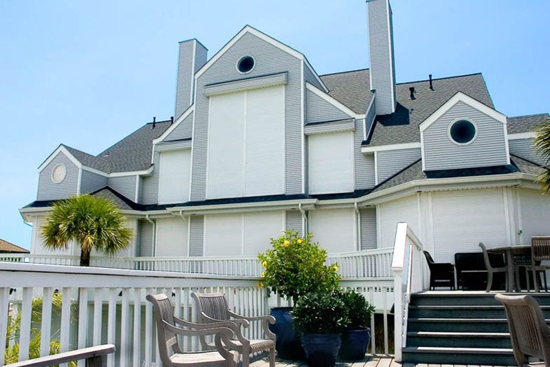 Coastal house with closed white hurricane shutters, viewed from a wooden deck with potted plants and chairs.