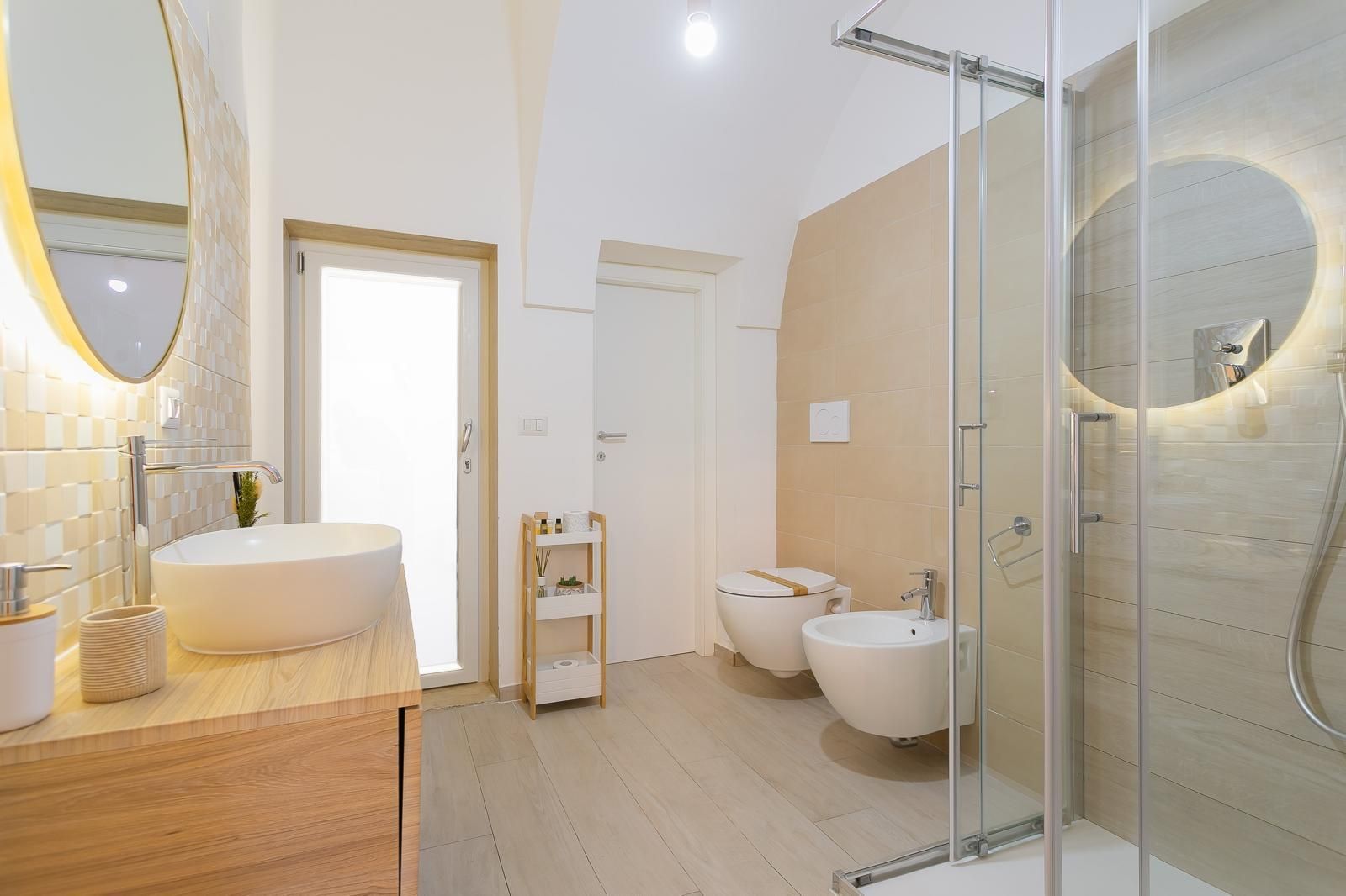 Bagno luminoso con doccia in vetro, WC, bidet, lavandino con mobile in legno e specchio illuminato.