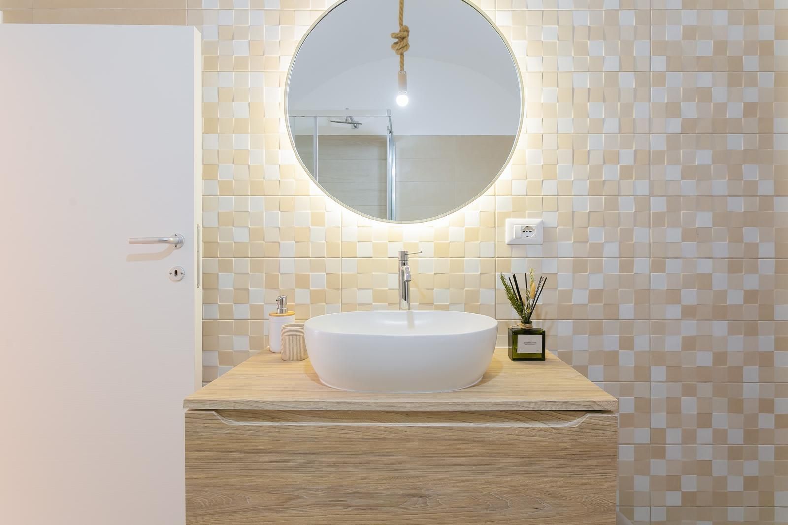 Bagno con lavandino bianco, specchio rotondo con luci e mobiletto in legno contro la parete piastrellata.