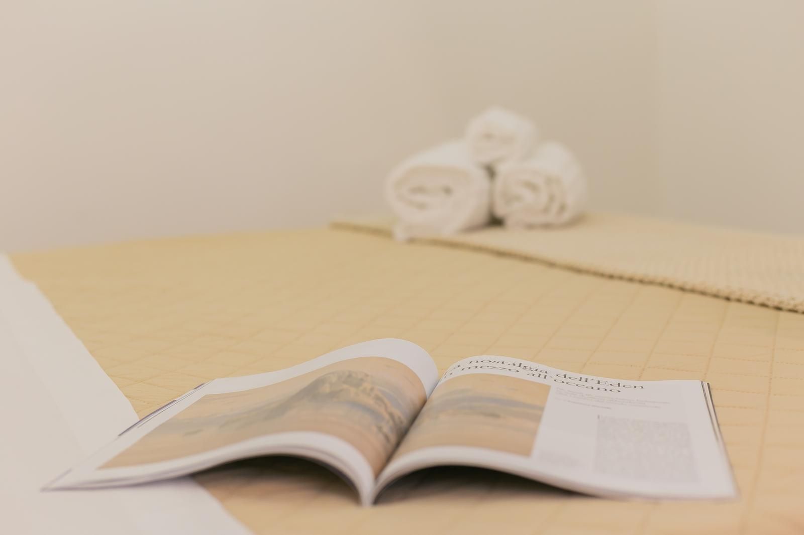 Una rivista aperta su un letto con asciugamani bianchi arrotolati sullo sfondo.