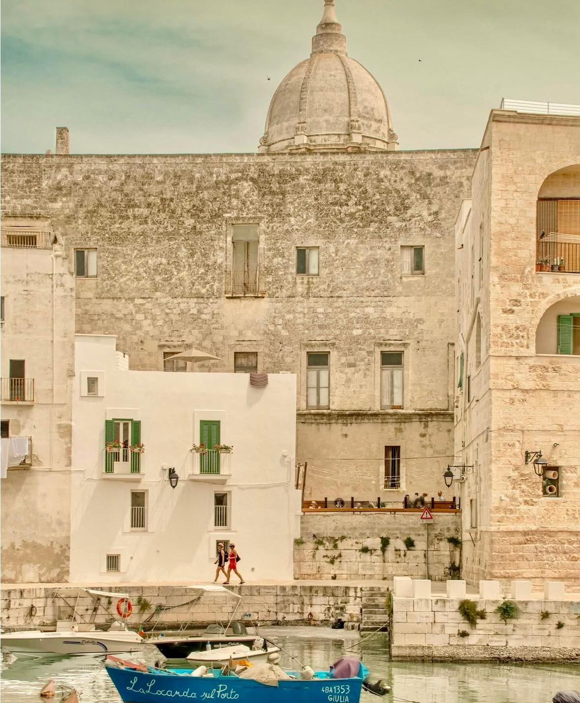 Edifici che si affacciano su un porto. Coppie che passeggiano lungo l'acqua. Edifici bianchi e beige con una struttura a cupola. Barca blu.