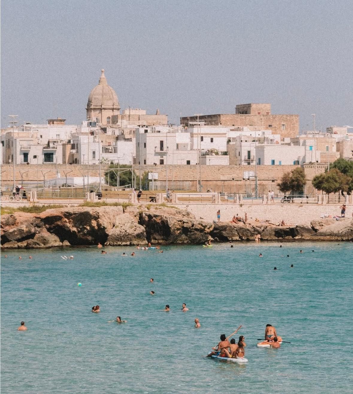 Città costiera con edifici bianchi e una chiesa con cupola. La gente nuota nelle acque turchesi.