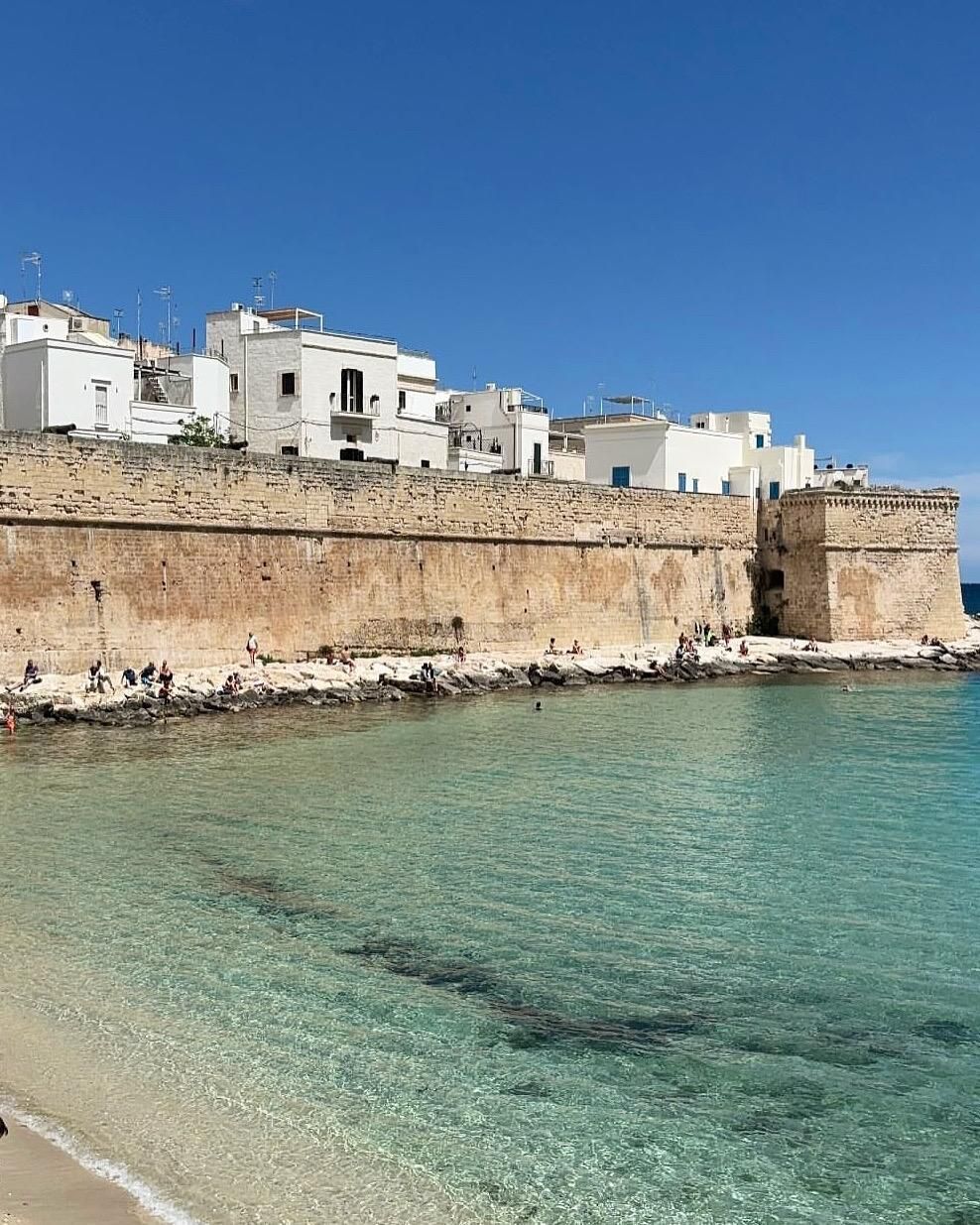 Edifici bianchi su una scogliera che domina un mare turchese con un muro di pietra e un cielo limpido.