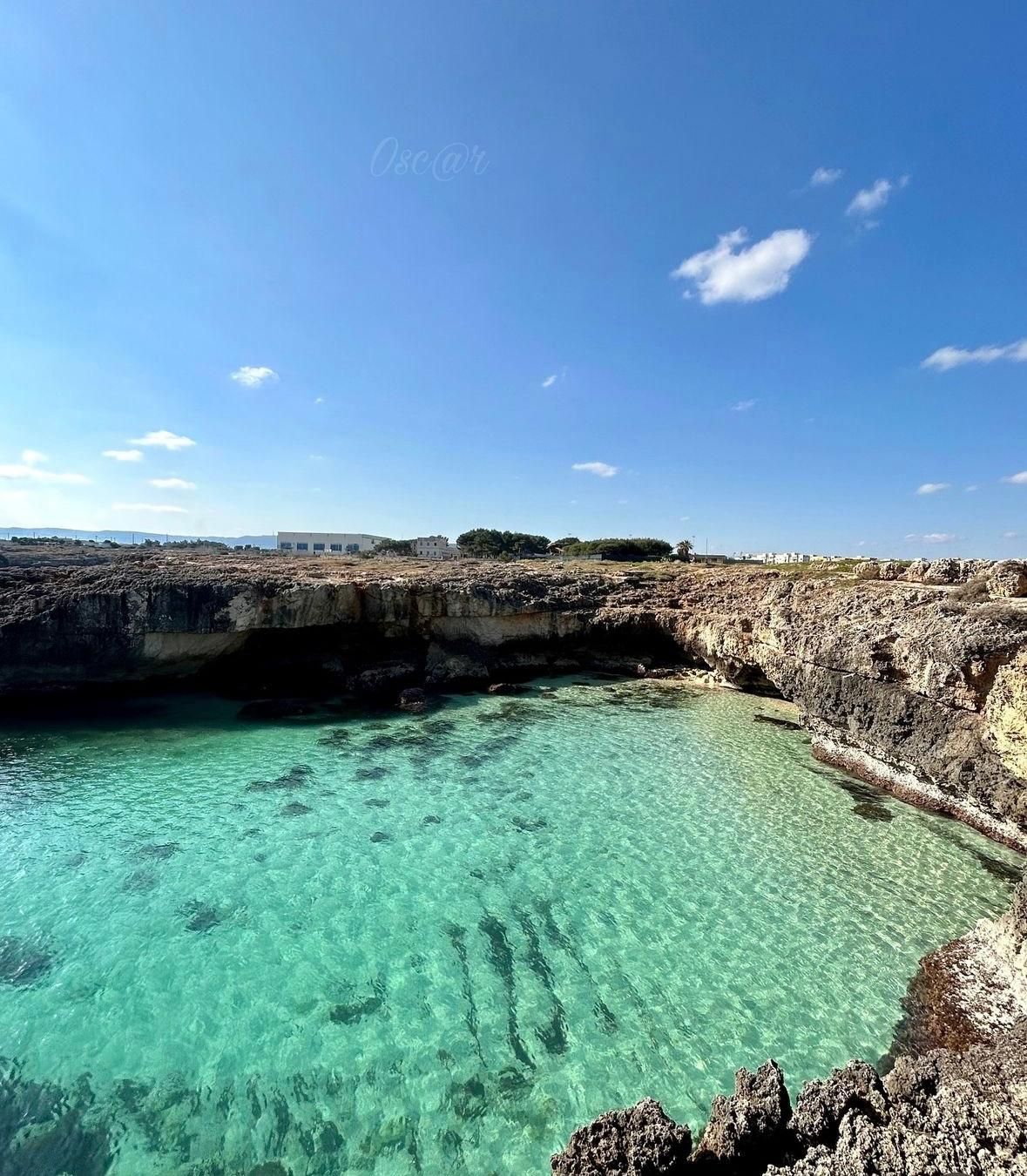 Acqua turchese in una baia rocciosa sotto un cielo azzurro. Edifici e alberi delineano l'orizzonte.