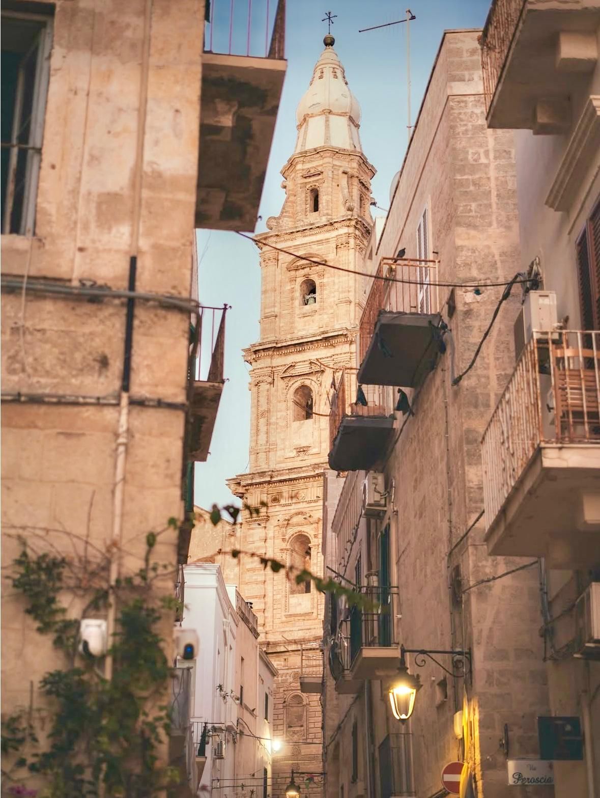Un alto campanile si erge tra vecchi edifici in una stretta strada italiana, sotto un cielo limpido.