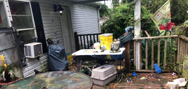 A cluttered outdoor deck with a table, bench, and plants; the backdrop is a white wall.