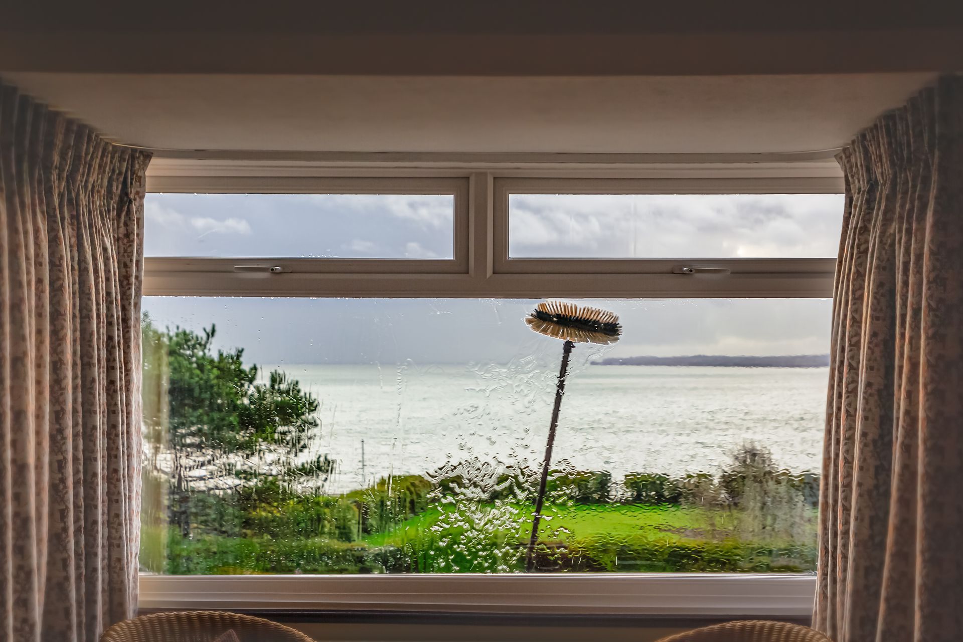 Window being cleaned with sea view, cloudy sky, green grass, and trees.