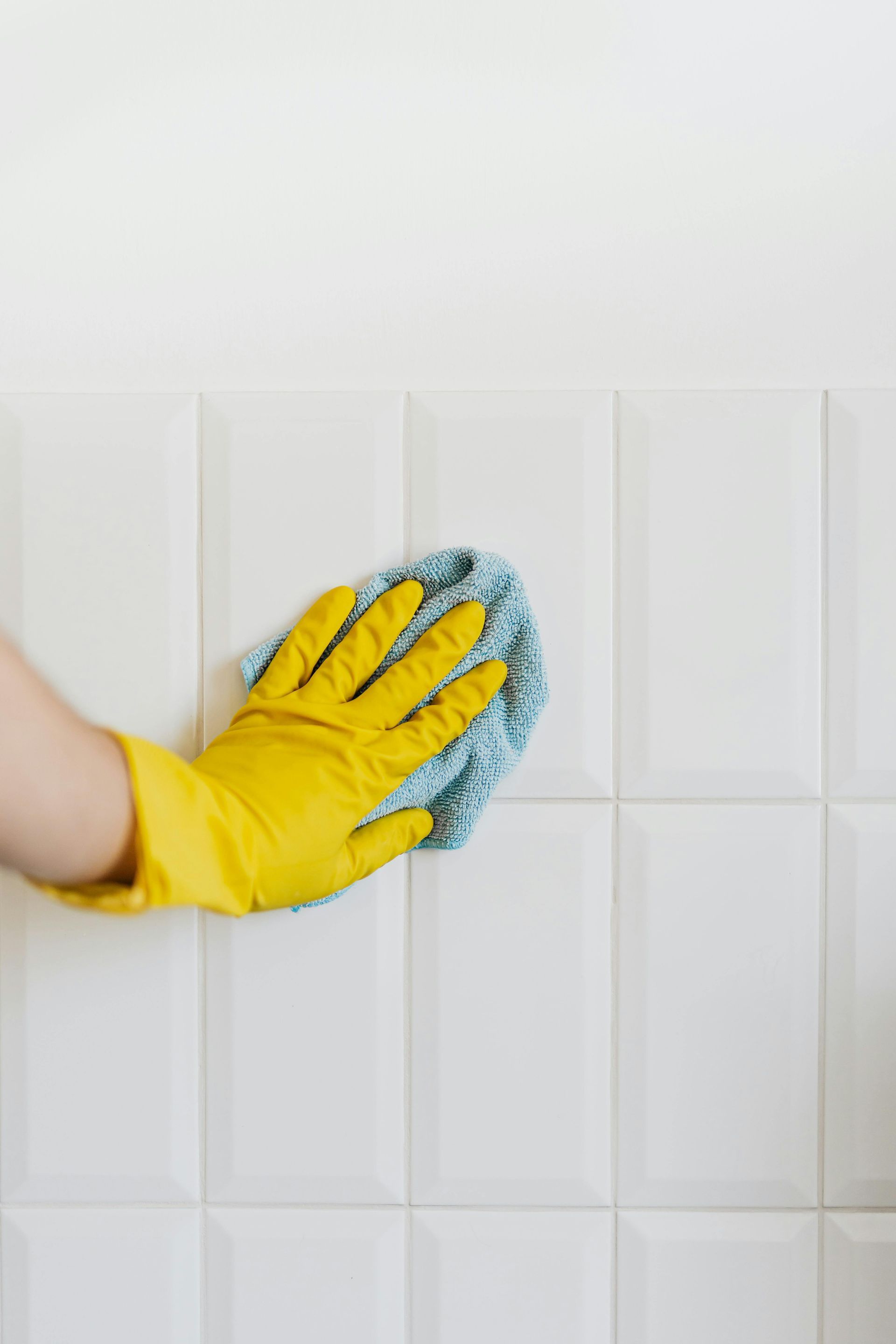 Hand in yellow glove wiping white tiled wall with blue cloth.