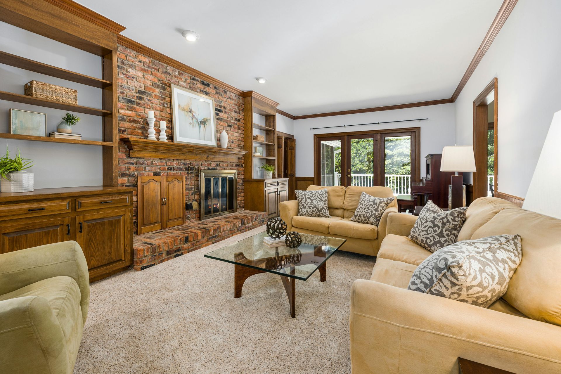 A living room with beige sofas, a glass-topped coffee table, a brick fireplace, and built-in wooden shelving.