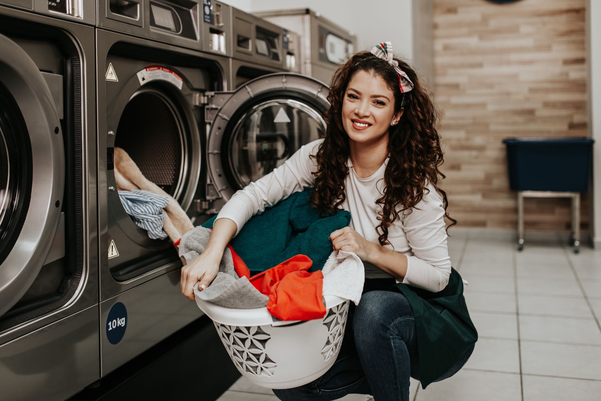 Woman loading laundry into a washer in a laundromat, smiling beside a basket of clothes.