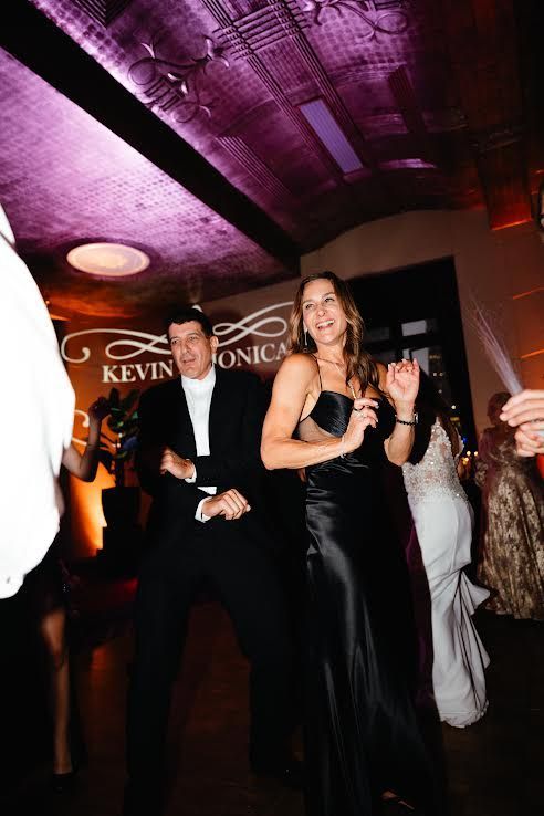 Couple dancing at a formal event, man in a tuxedo, woman in a black dress, logo on the wall, warm lighting.