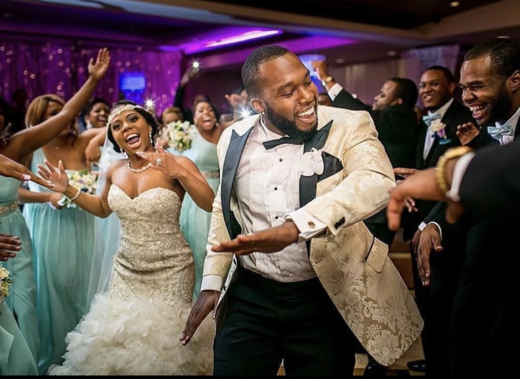 Newlyweds and wedding party dancing, celebrating joyfully in a reception hall.