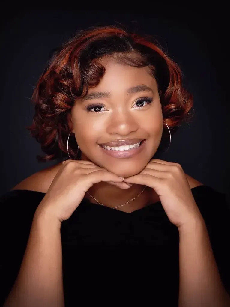 A young woman with red hair is smiling with her hands on her chin.