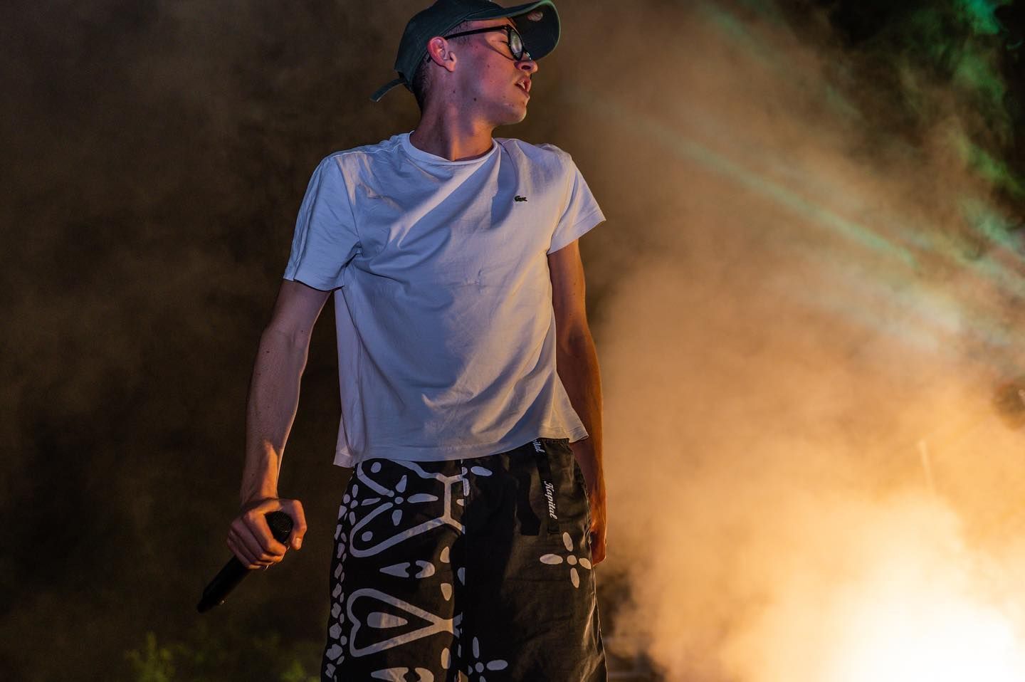 Rapper sul palco, guarda a sinistra, con il microfono in mano. Indossa un berretto, occhiali, una maglietta bianca e pantaloni a fantasia. Fumo e luci sullo sfondo.