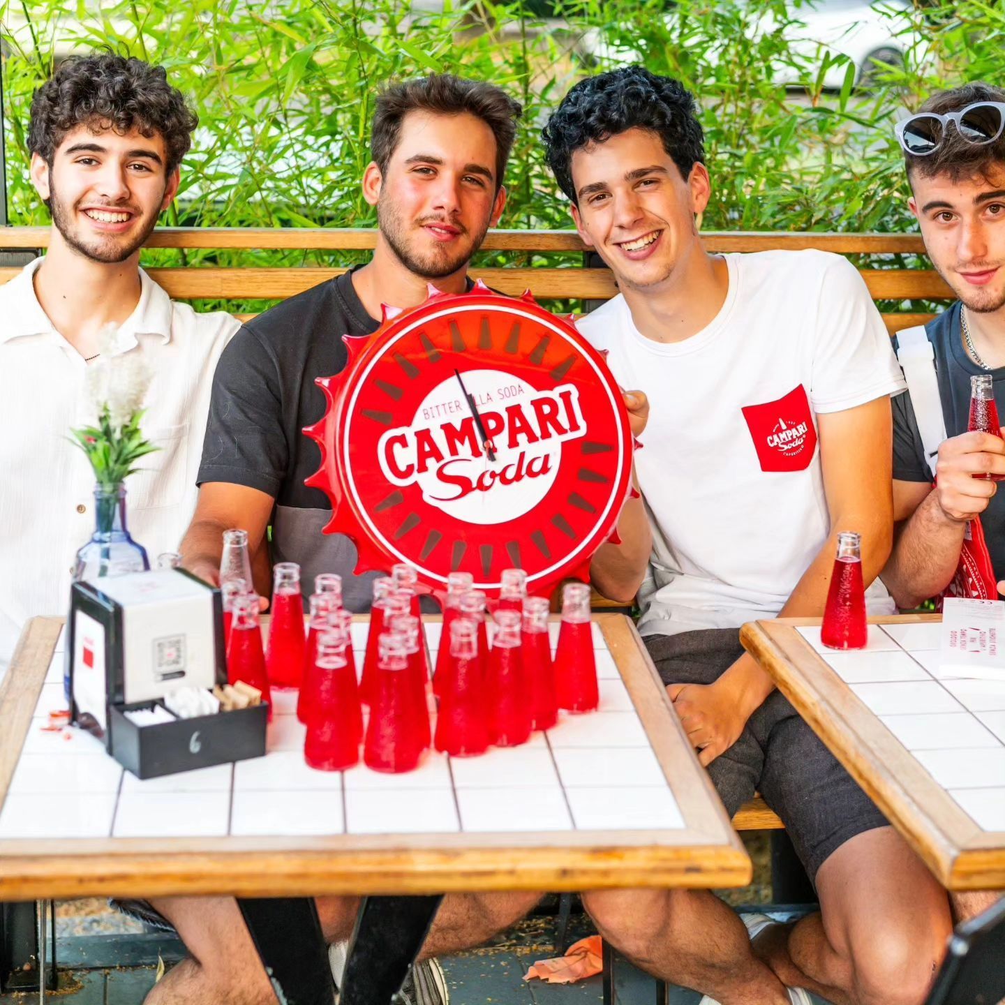 Quattro giovani seduti a un tavolo con bottiglie di Campari Soda, sorridenti, tengono in mano un cartello con la scritta Campari Soda.