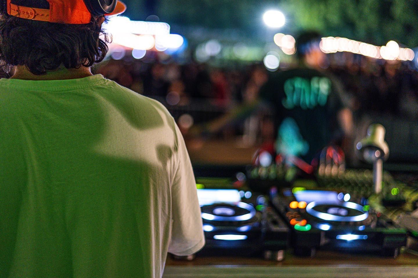 Due DJ a un evento all'aperto. Uno in verde, l'altro con la maglietta dello staff, con l'attrezzatura e una folla sfocata.