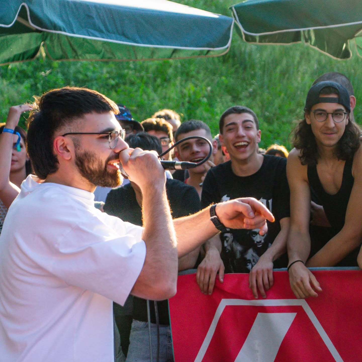 Un uomo con la barba e gli occhiali canta in un microfono, indicando la folla.