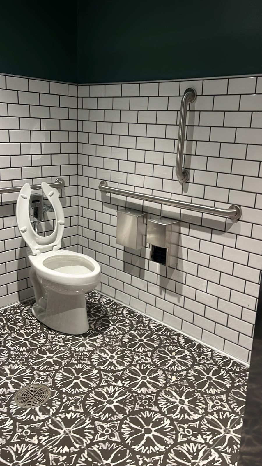 A public restroom with white brick-style tiled walls, patterned floor, and accessible grab bars.