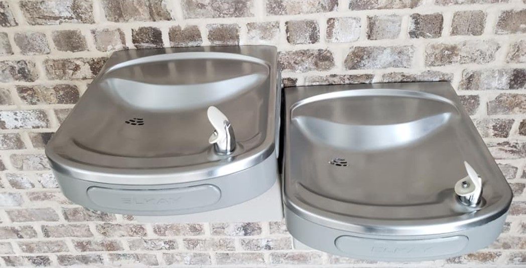 Two stainless steel water fountains on a brick wall. One is slightly darker than the other.