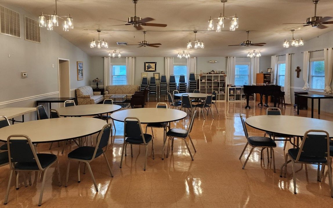Large community hall with round tables, chairs, and a piano. Light-colored walls and ceiling fans.