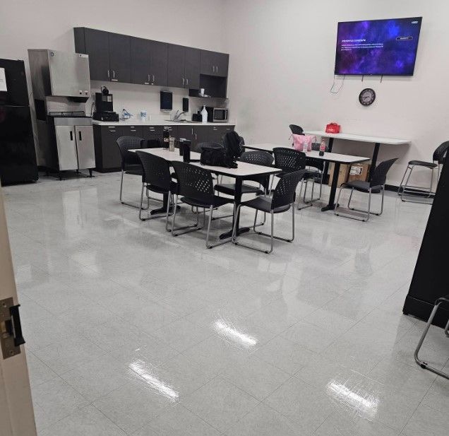 An empty break room with tables, chairs, a kitchenette with cabinets, and a television on the wall.
