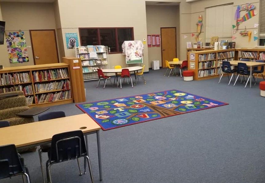 School library with bookshelves, tables, and a colorful tree-themed rug.