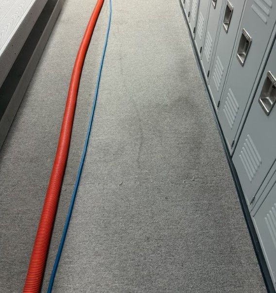 A long, gray carpeted hallway with lockers; a red and blue hose lie across it, as if cleaning.