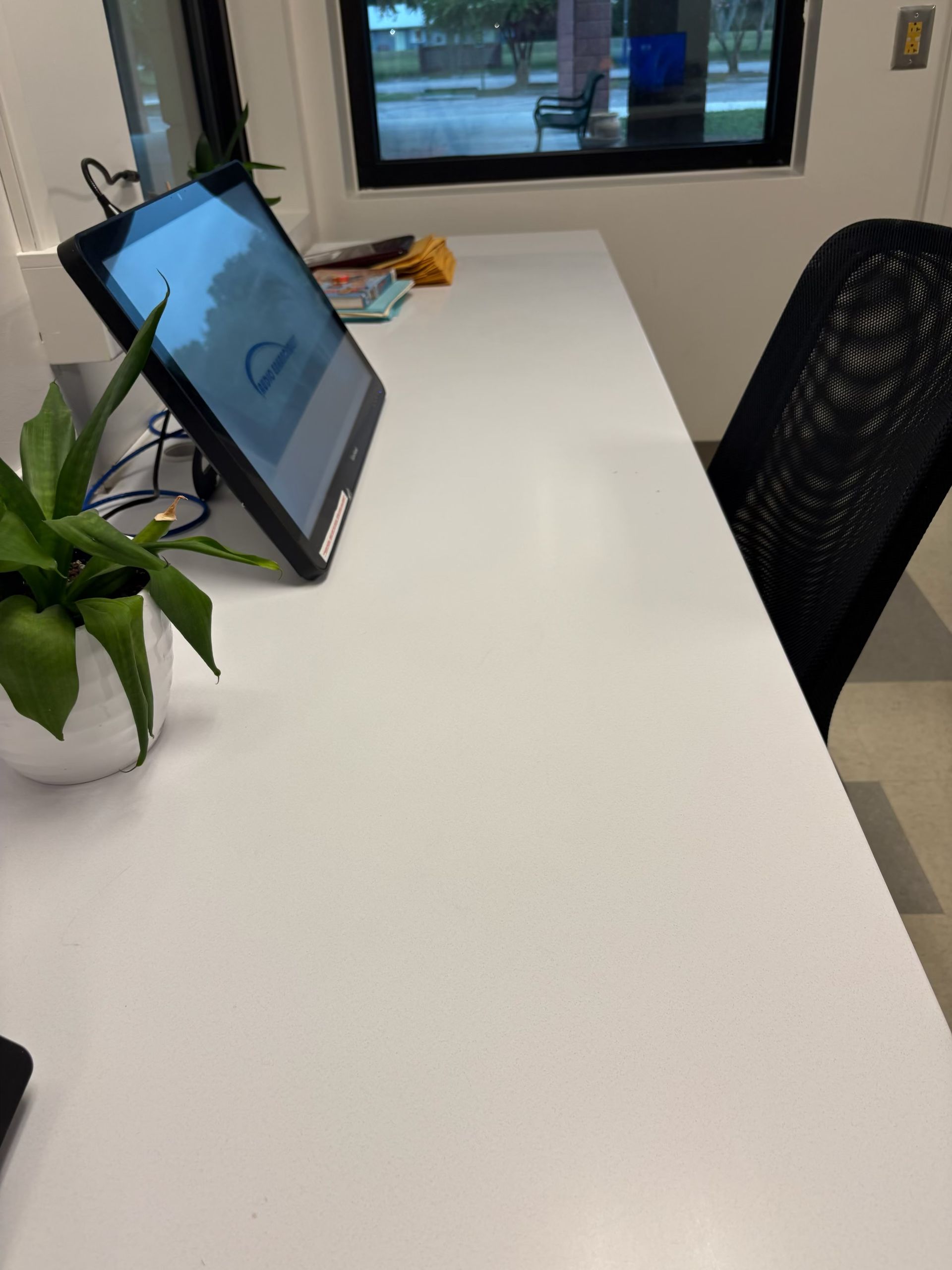 White desk with a computer monitor, a plant, and a black chair in front of a window.
