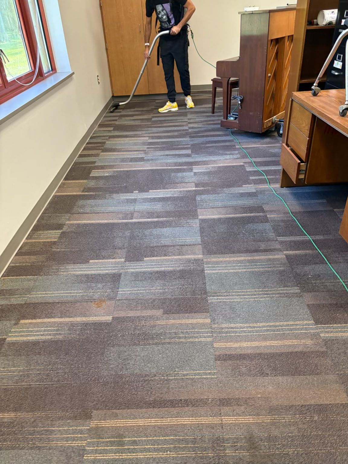 Person vacuuming carpet in a room with a piano and desk.