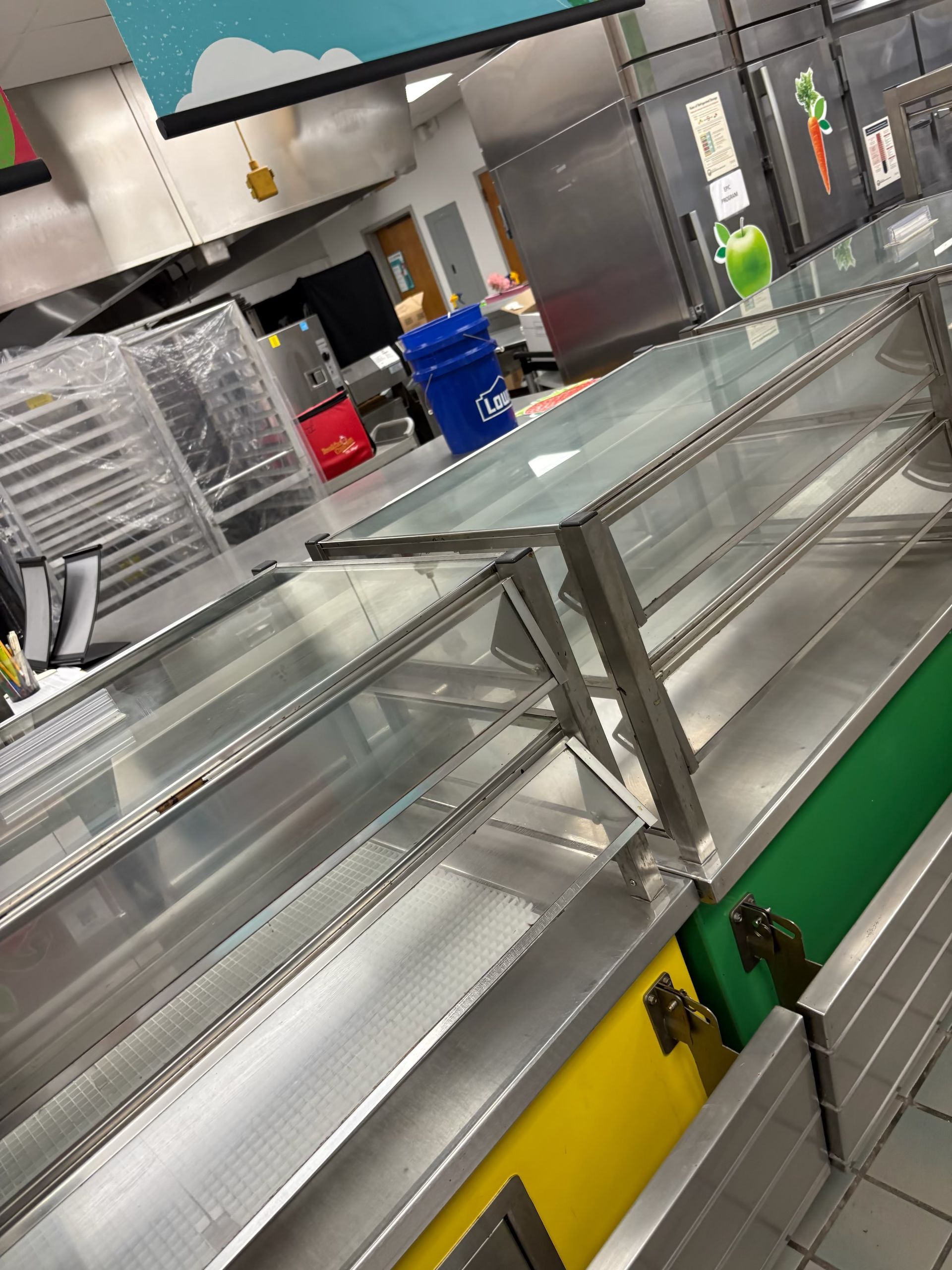 Empty cafeteria serving line; stainless steel with yellow and green panels; overhead signage.