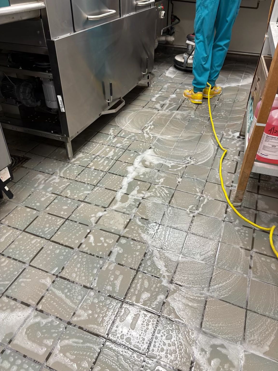 Person in blue scrubs and yellow boots cleaning tiled floor with a machine. Soap and water visible.