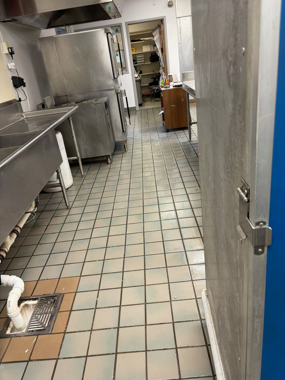 A commercial kitchen with stainless steel appliances and tiled floor. A door is on the right.